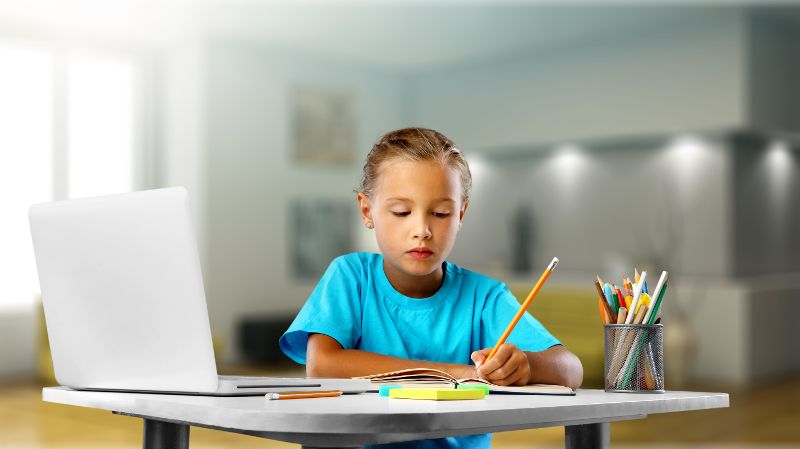 Ein kleiner Junge schreibt an einem Tisch sitzend etwas in sein Heft. Daneben steht ein Laptop und weitere Stift in einem Behälter.