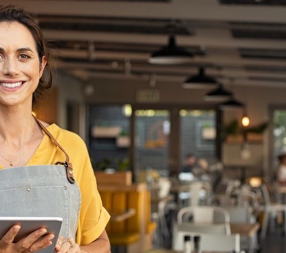 Eine Frau mit Schürze bekleidet steht mit einem Tablet in der Hand am linken Bildrand, im Hintergrund ist ein Restaurant.